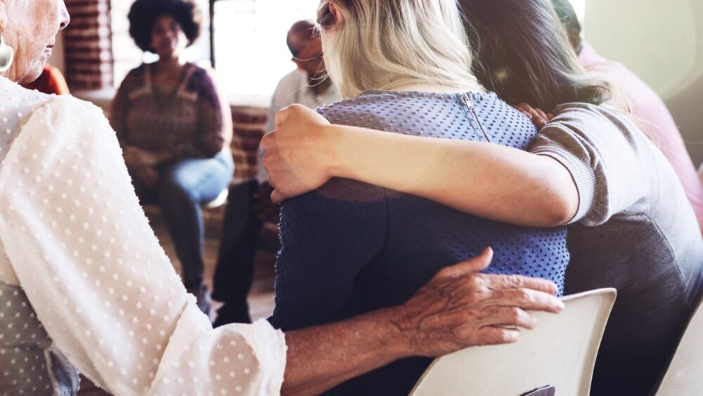 Support group meeting with individuals sitting in a circle hugging and supporting one another.
