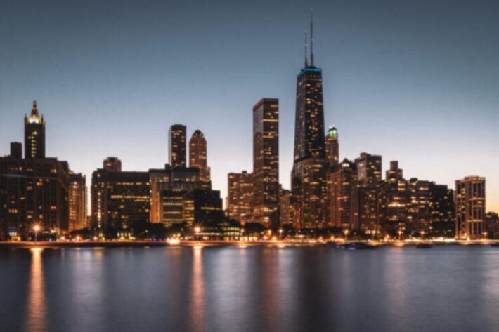 Chicago skyline at dusk with illuminated buildings reflecting on the calm water, featuring the John Hancock Center.