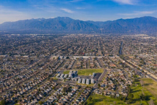 Aerial view of a suburban neighborhood with a grid-like pattern of streets and houses, surrounded by green spaces and a school campus in the foreground. The background features a mountain range under a clear blue sky, with a long road cutting through the cityscape.