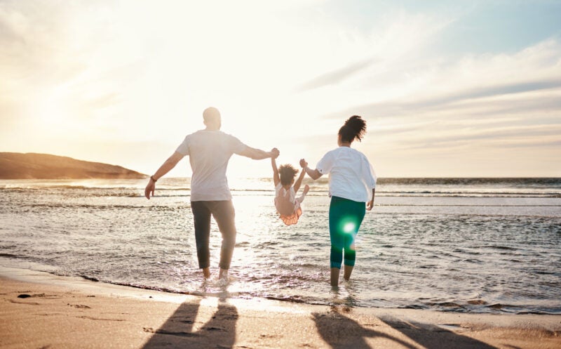 Dad, mom swing their daughter while walking on the beach.