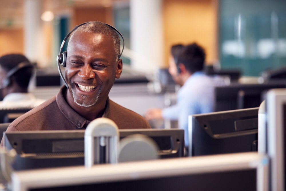 A smiling customer service representative wearing a headset in a call center, surrounded by computer monitors and colleagues working in the background.
