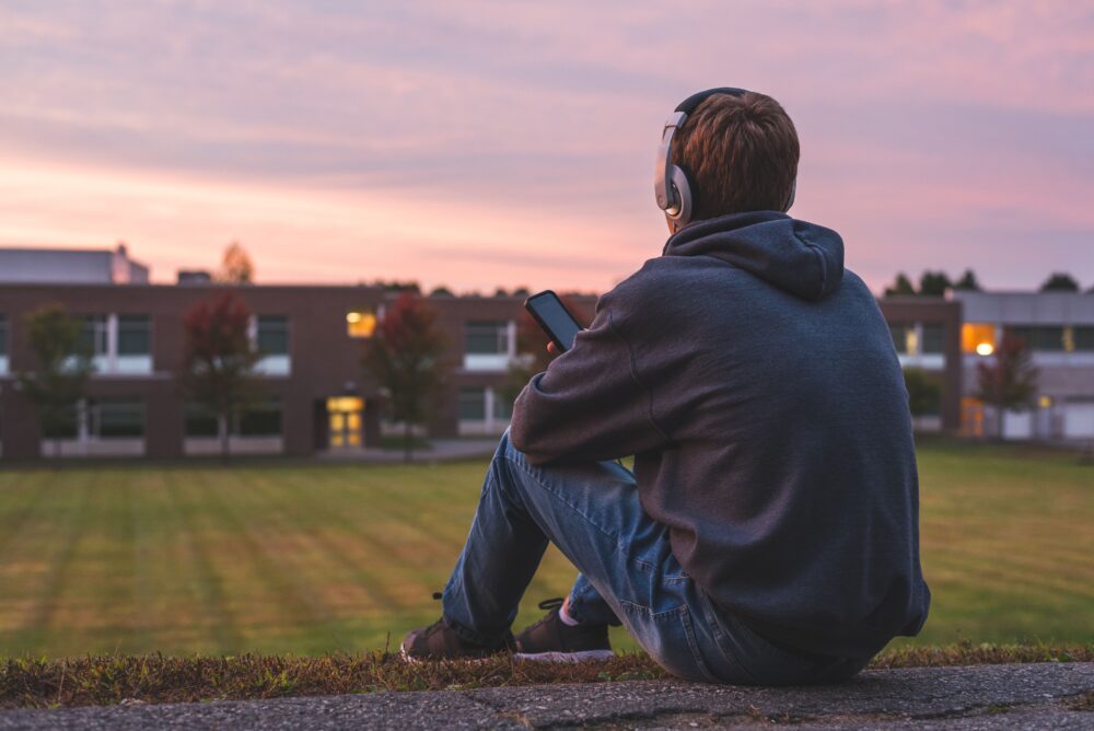 A teen sits on a hill overlooking a school, wearing headphones.