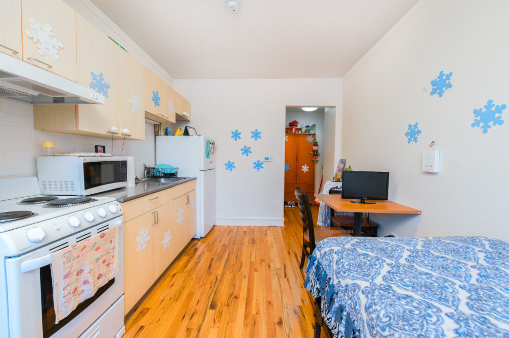 Photo of the interior of a Permanent Supportive Housing unit. Image shows a bed and kitchen with bright lighting.