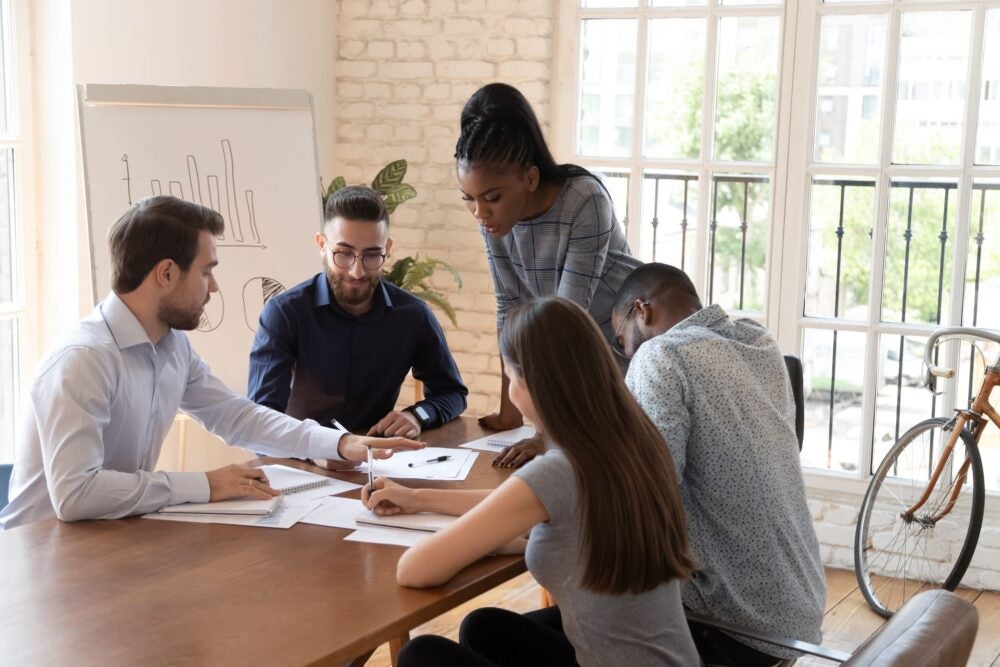 A group of five colleagues gathered around a table in a bright office, collaborating on documents with charts on a whiteboard in the background.