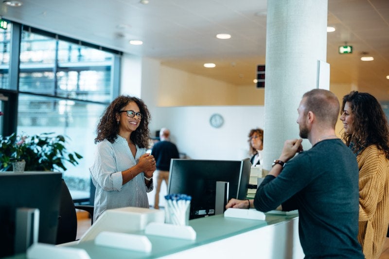 A receptionist smiles and speaks with two visitors standing at a front desk in a bright, modern office lobby.