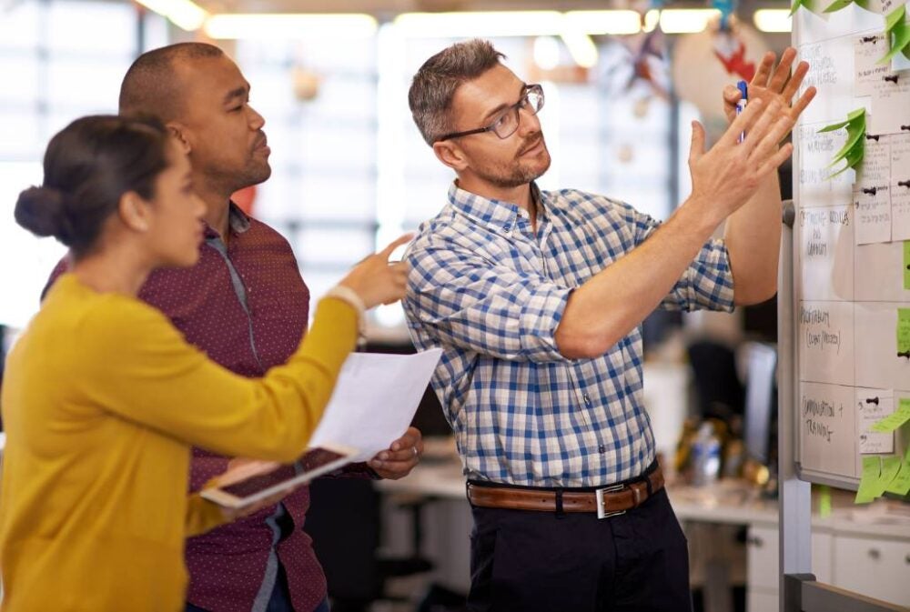 Three coworkers reviewing notes on a whiteboard in an office.