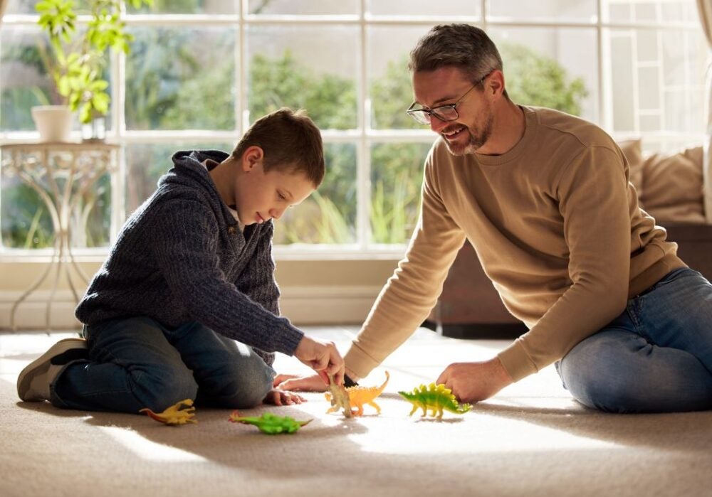 A father and son play with toy dinosaurs on the floor.
